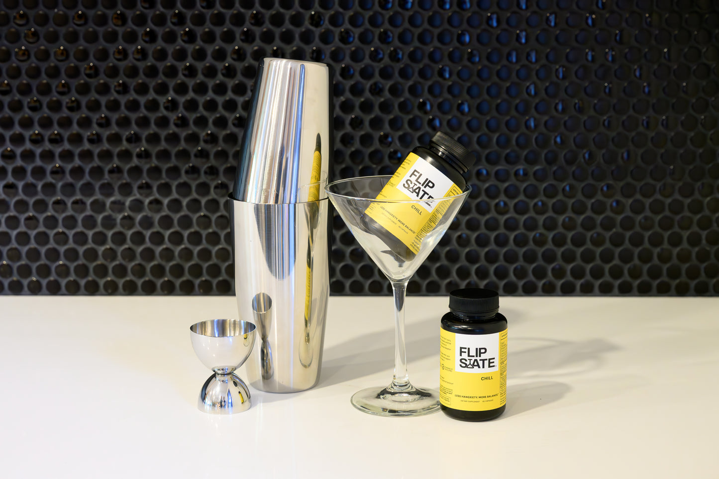 Bartending tools including a cocktail shaker, jigger, and bottles of FlipSalte on a textured black background.