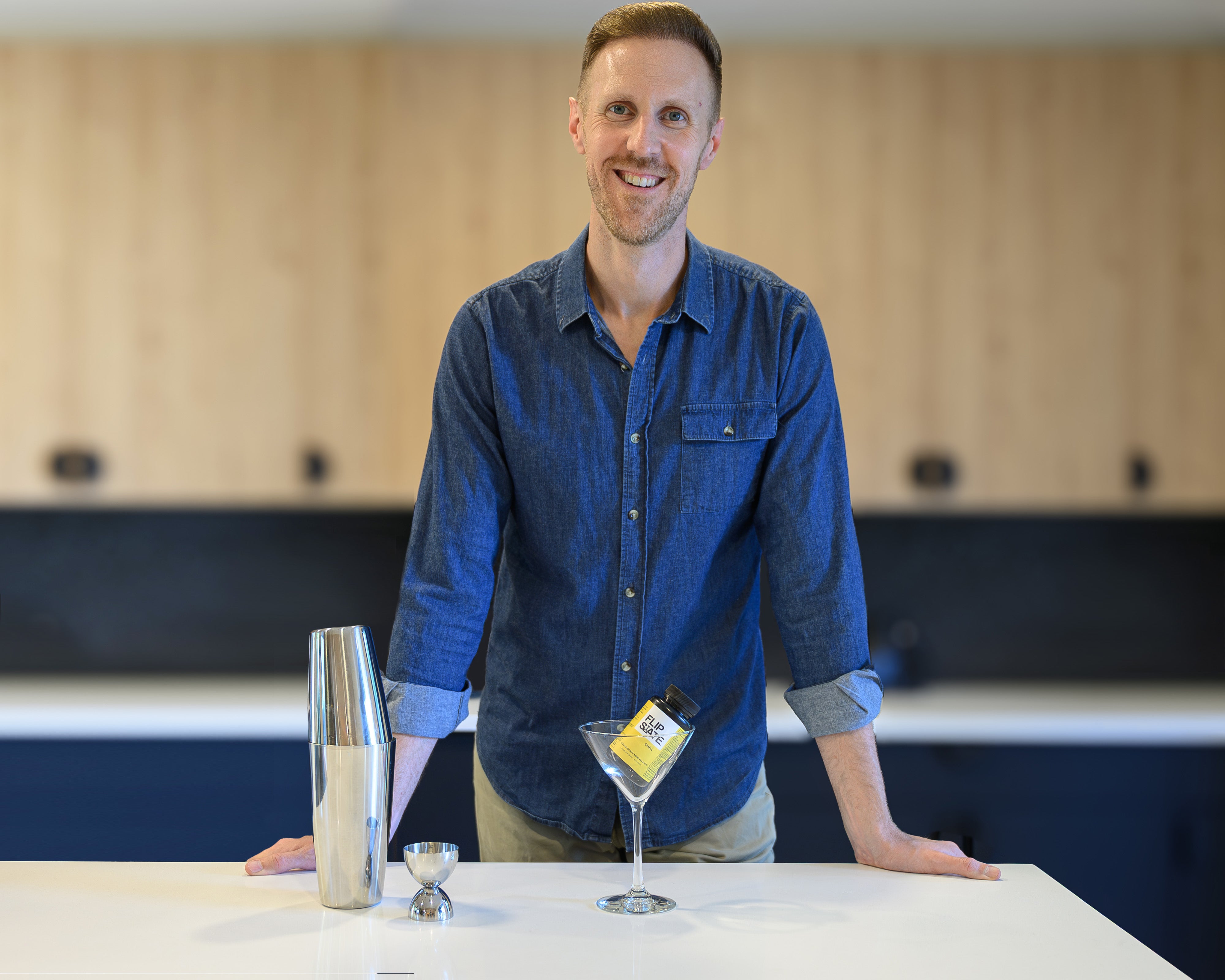 Man in a blue shirt standing behind a bar with a cocktail shaker and glass containing bottle labeled 'FlipState' which is a hangxiety relief supplement